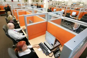 Wielostanowiskowe biuro, gdzie pracownicy obsługują telefony i komputery. W tle widać wiele przegródek, co może sugerować pra group nękanie.