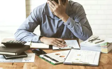 Zestaw dokumentów, kalkulator i telefon leżą na biurku. Mężczyzna z ręką na czole, przytłoczony problemami finansowymi, przegląda pismo windykacyjne.